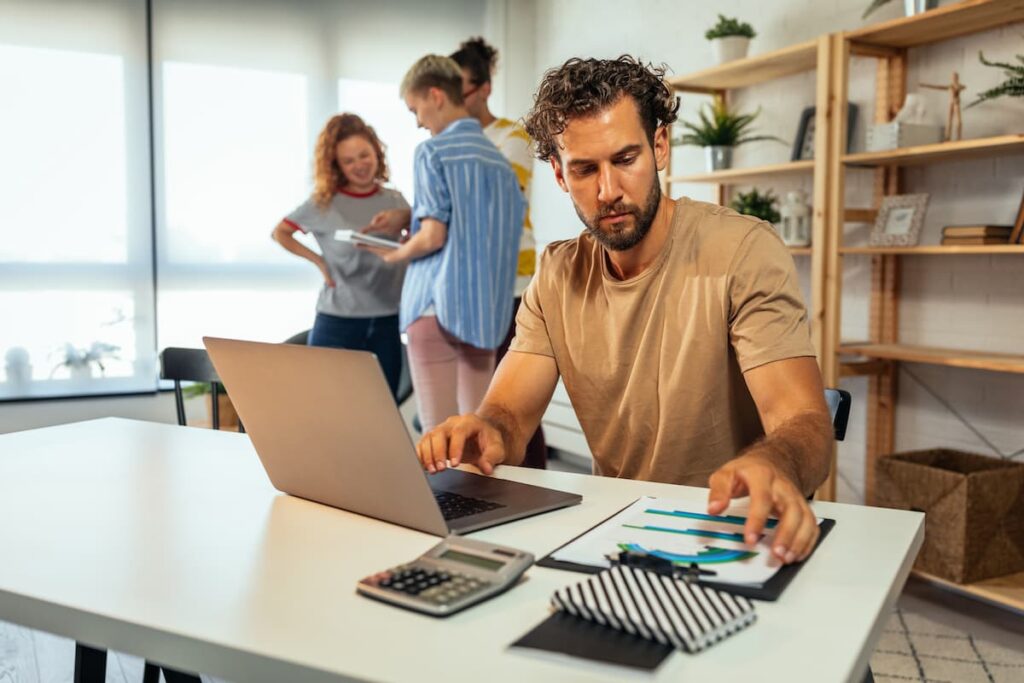 En mand som sidder og strukturere sit arbejde på et skrivebord med mennesker i baggrunden som har en dialog